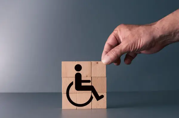Simboli di accessibilità su cubi di legno, con una mano che aggiunge l'ultimo pezzo. Rappresenta il **certificato medico introduttivo per accompagnamento**.