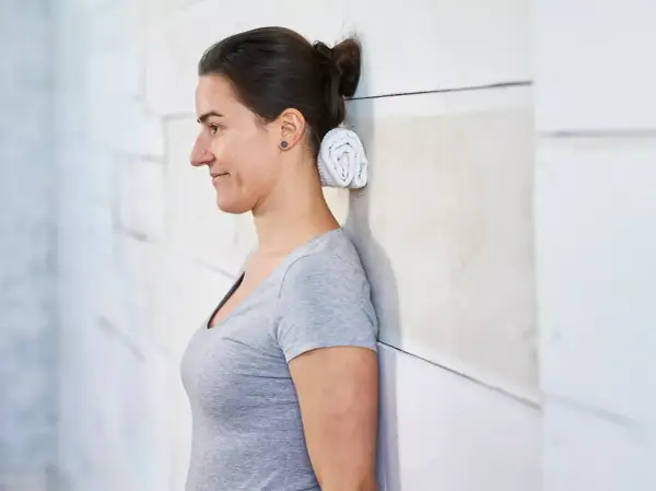 Donna con asciugamano arrotolato dietro il collo, appoggiata a un muro. Potrebbe alleviare il mal di testa da cervicale.