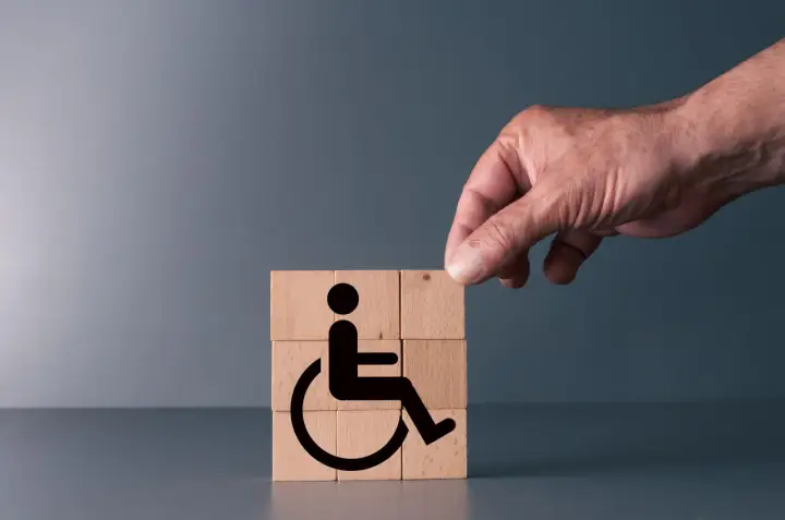 Simboli di accessibilità su cubi di legno, con una mano che aggiunge l'ultimo pezzo. Rappresenta il **certificato medico introduttivo per accompagnamento**.